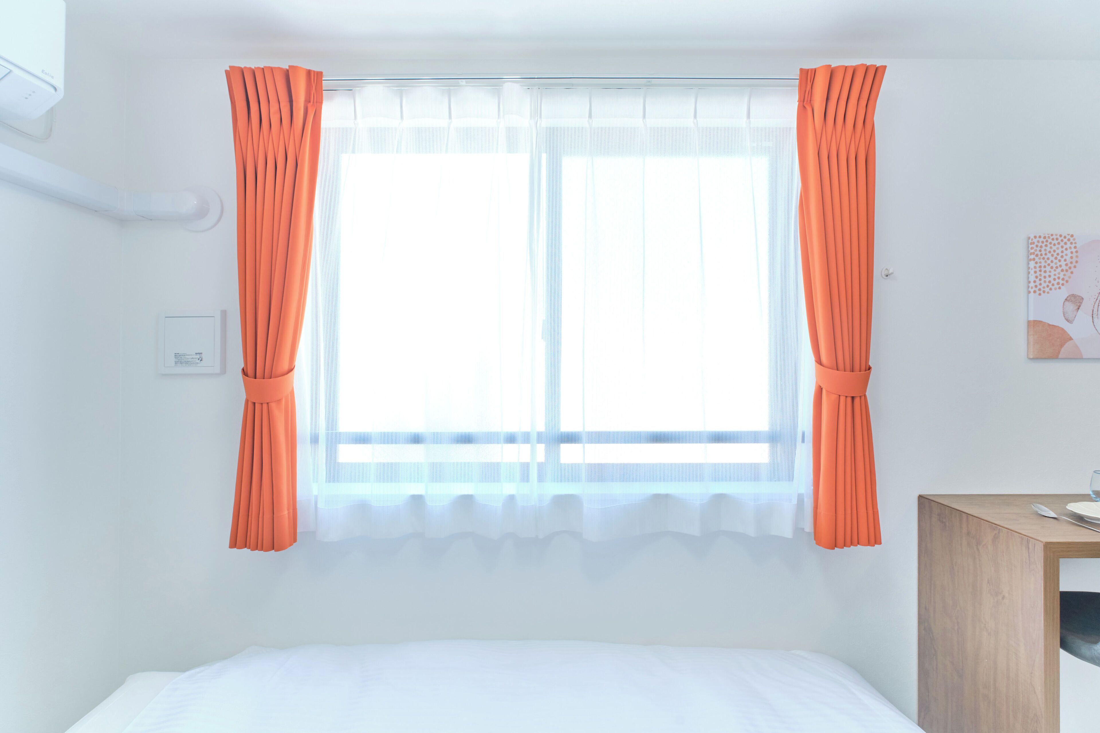 A room with a bed, a wooden side table with a glass, and a window with orange curtains.
