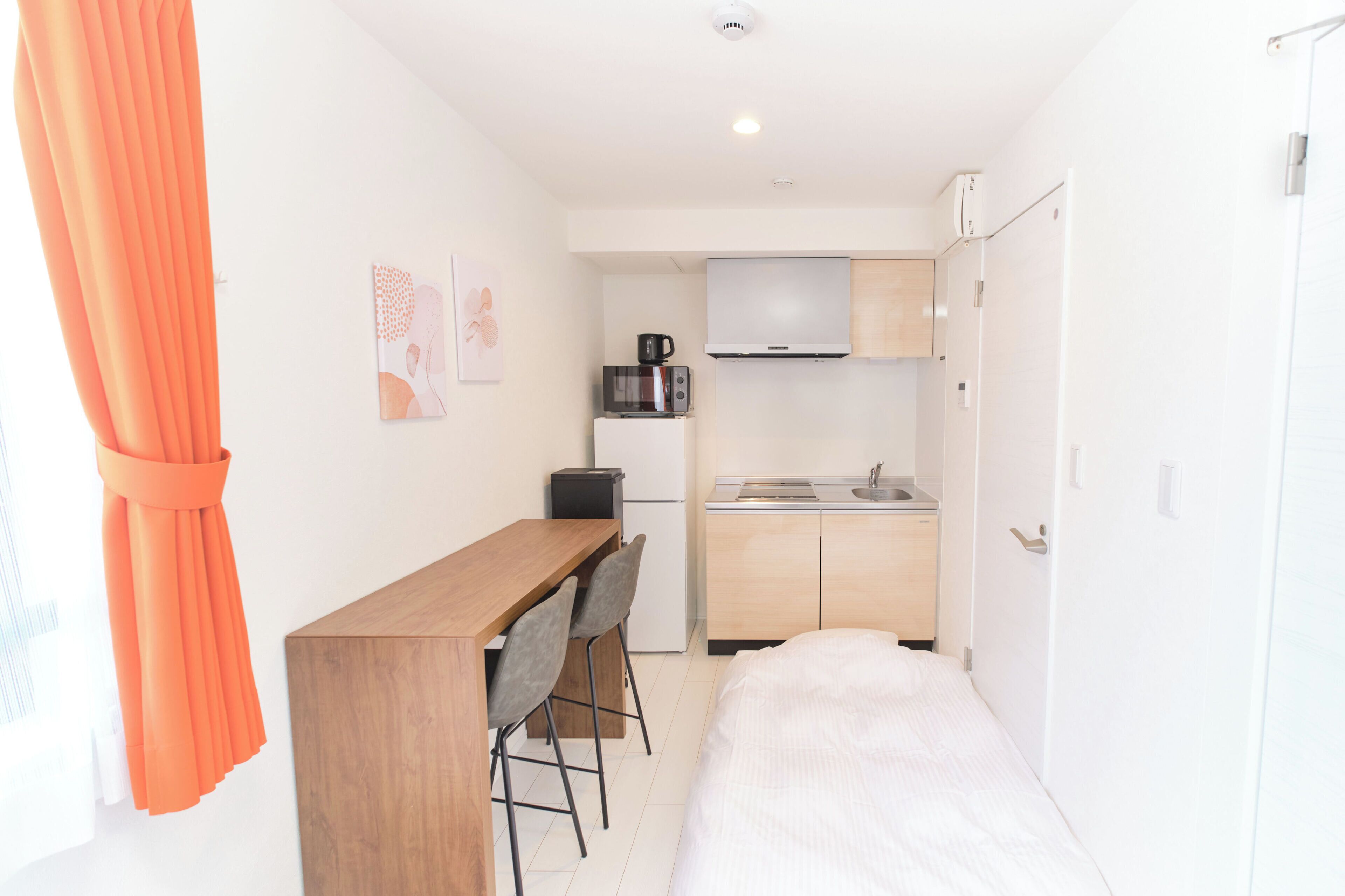 A compact room with a bed, a small kitchenette, a wooden desk with two chairs, and a window with orange curtains.