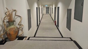 Un pasillo largo y estrecho con puertas a ambos lados, un extintor a la derecha y un jarrón decorativo a la izquierda.