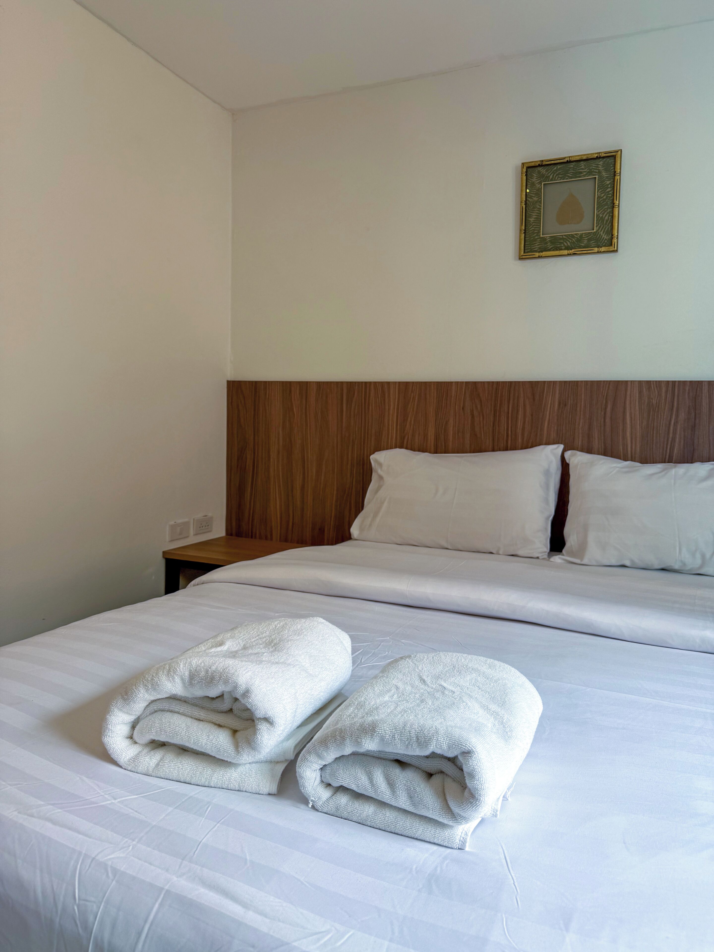 A hotel room with a neatly made bed, a wooden headboard, a framed picture on the wall, and a nightstand with a lamp.