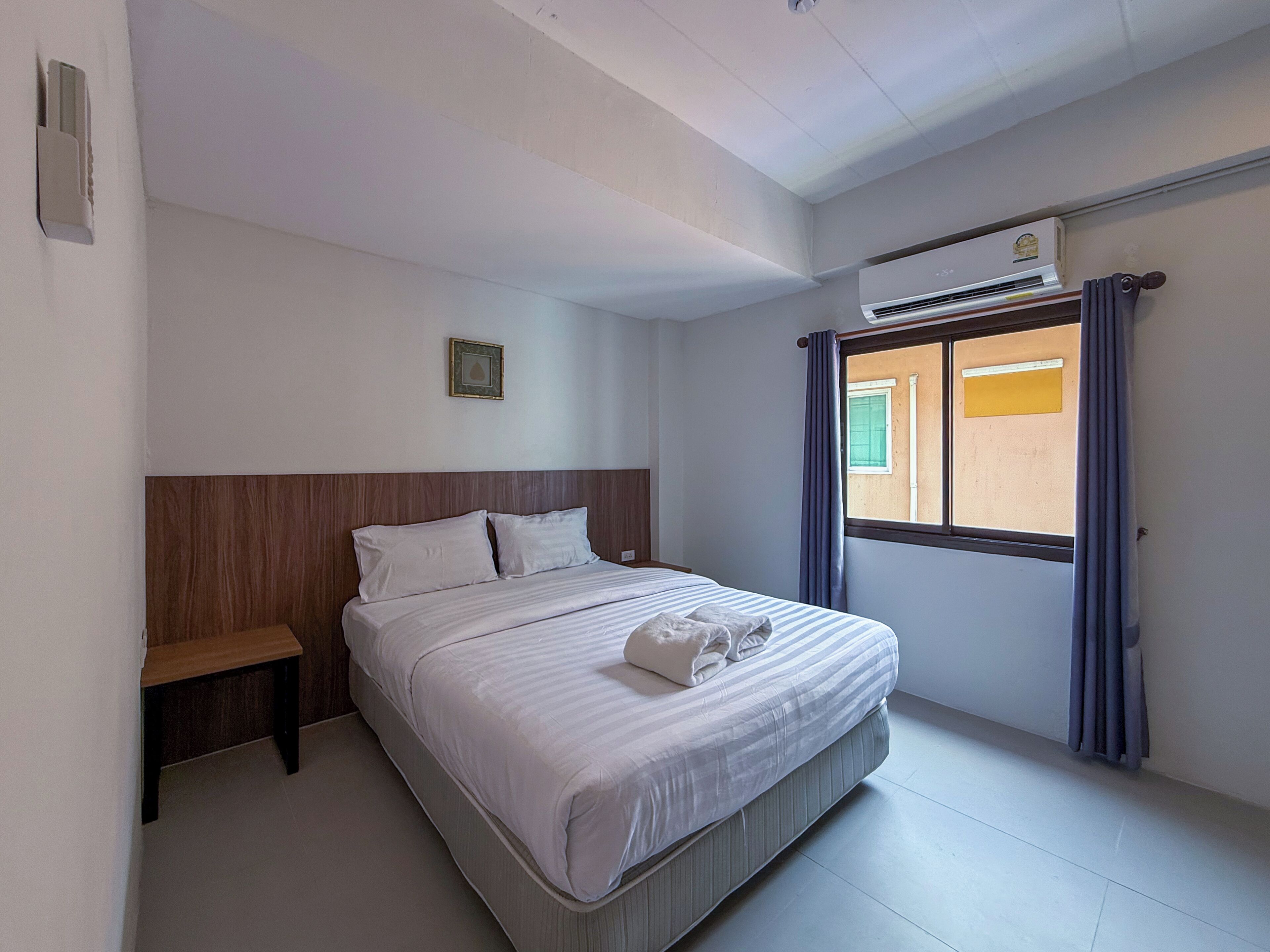 Ein Hotelzimmer mit Bett, Nachttisch, Fenster mit Vorhängen und einer Klimaanlage.