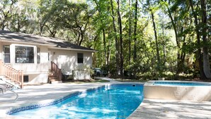 Una piscina con borde de azulejos, una casa con porche y un área boscosa al fondo.