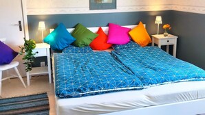 Ein Schlafzimmer mit einem Bett, farbenfrohen Kissen, einem Nachttisch mit Lampe, einem gerahmten Bild an der Wand und einer Pflanze.