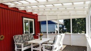 Eine überdachte Terrasse mit einem weißen Tisch und Stühlen, einer roten Wand und Blick auf ein blaues Haus.