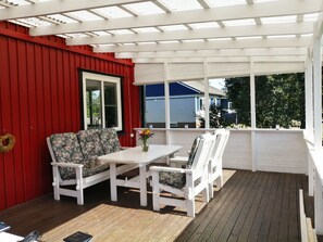 A covered patio with a white table and chairs, a red wall, and a view of a blue house. - Buntes Schwedenhaus Lillebror by Interhome (Fågelfors)