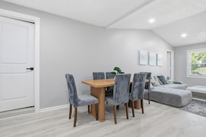 A dining area with a wooden table and grey upholstered chairs, a grey sofa, and a window with a view of greenery.