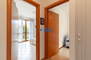 Ein Zimmer mit Holztür, einem Bett mit weißer Bettdecke, einem kleinen Tisch und einem Fenster mit Vorhängen.