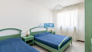 Un dormitorio con dos camas verdes, una mesita de noche con lámpara, una ventana con cortinas traslúcidas y un armario verde.
