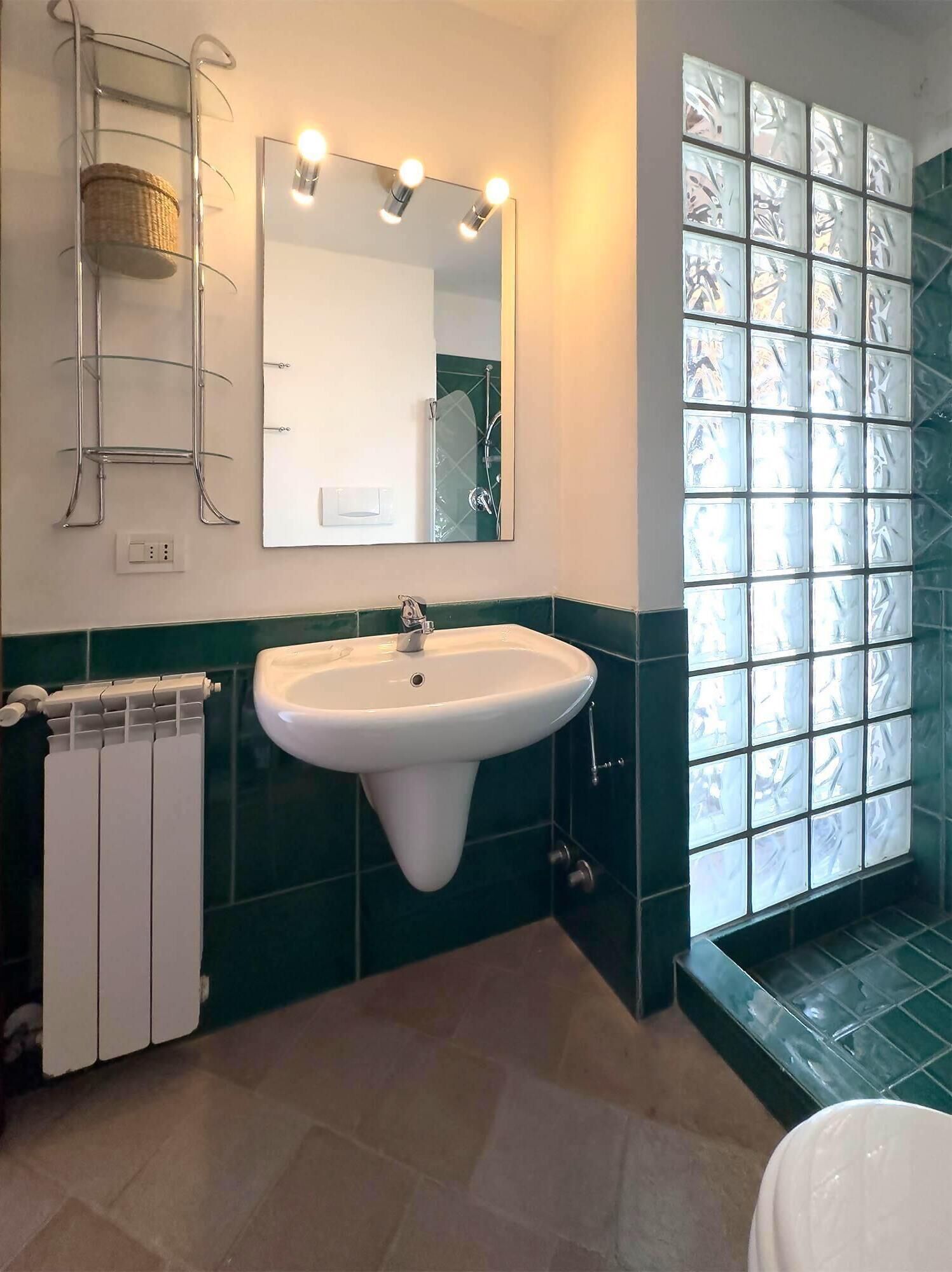 A bathroom with a white sink, a mirror with lights, a towel rack, and a green-tiled wall.