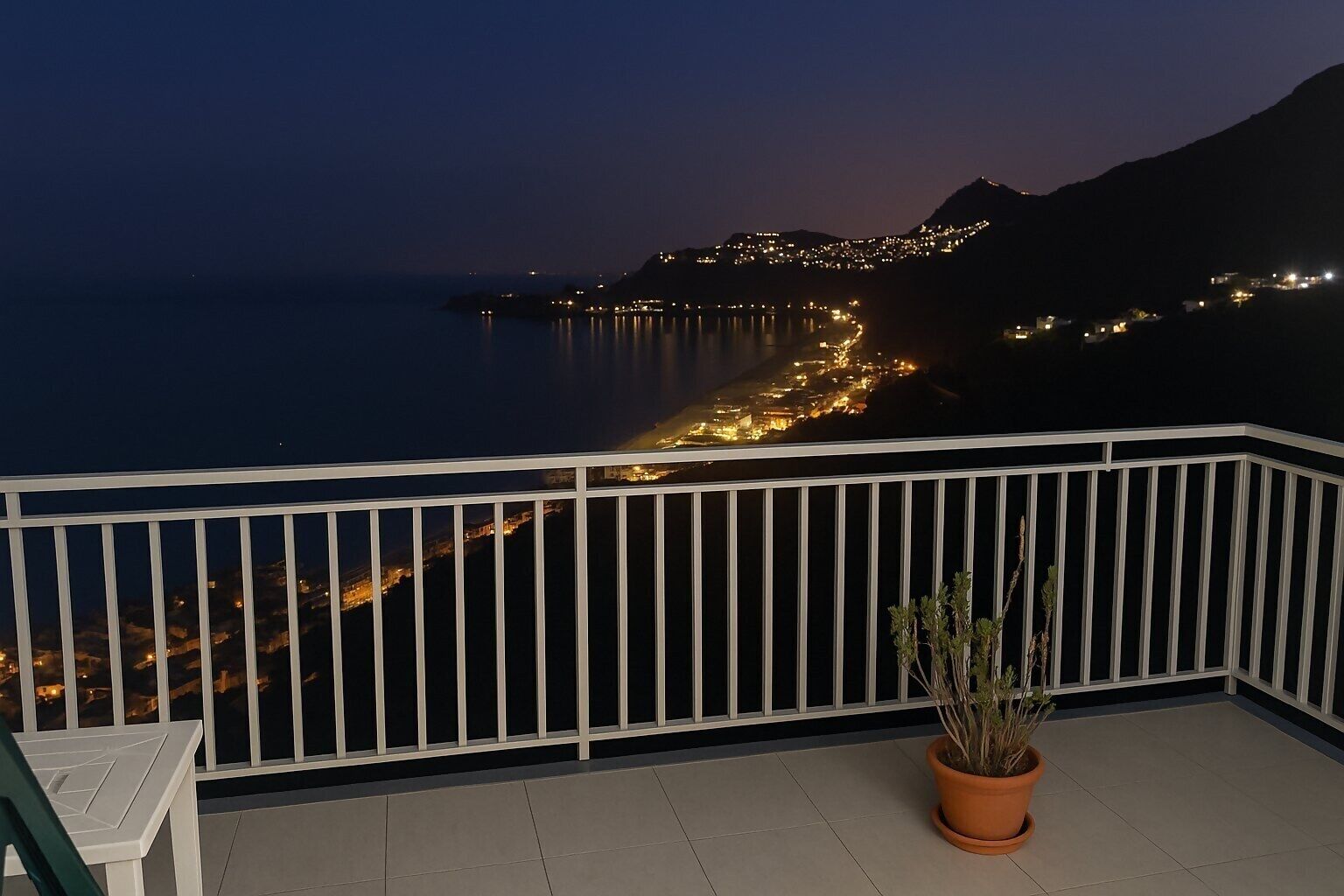 Un balcón con barandilla, una planta en maceta y vistas de un pueblo costero de noche.