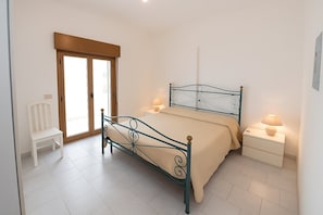 Ein Schlafzimmer mit einem Metallbettgestell, einem weißen Stuhl, einem Nachttisch mit Lampe und einer Schiebetür aus Glas.