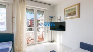 Un salon moderne avec une télévision, un canapé, un fauteuil, un balcon offrant une vue sur les immeubles et un panneau indiquant « Aperta Solo A Ribalta ».