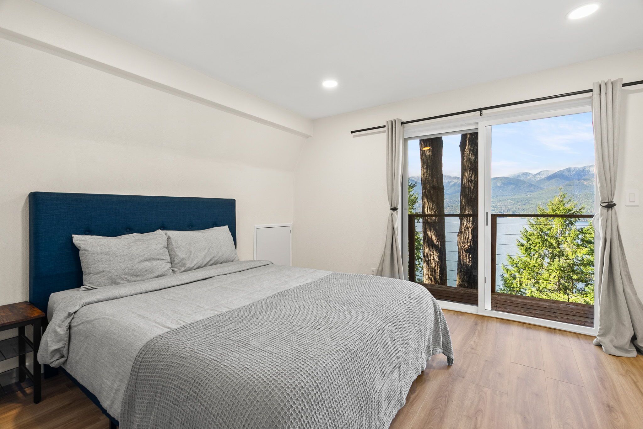 A bedroom with a bed, a nightstand, a large window with curtains, and a view of mountains.