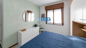Ein Schlafzimmer mit Bett, Kommode, Stuhl, Fenster mit Vorhängen und einem Spiegel an der Wand.