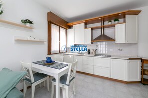 A modern kitchen with white cabinetry, wooden shelves, and a dining area with a table and chairs. - ° Emily °- [three-room apartment] #200m from the sea# (Jesolo)
