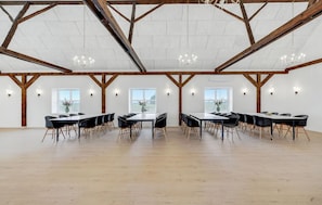 Ein geräumiger Saal mit hohen Decken, Holzbalken und Kronleuchtern. Tische und Stühle sind für Essen oder Besprechungen aufgestellt.
