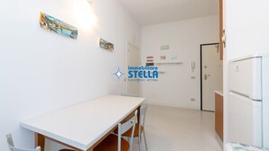 Ett litet, rent rum med ett vitt bord, två stolar, ett kylskåp och en dörr som leder till ett annat rum.