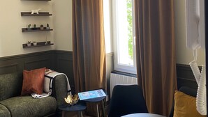 Een woonkamer met een bank, een bijzettafeltje, een stoel, een raam met gordijnen en planken met decoratieve objecten.
