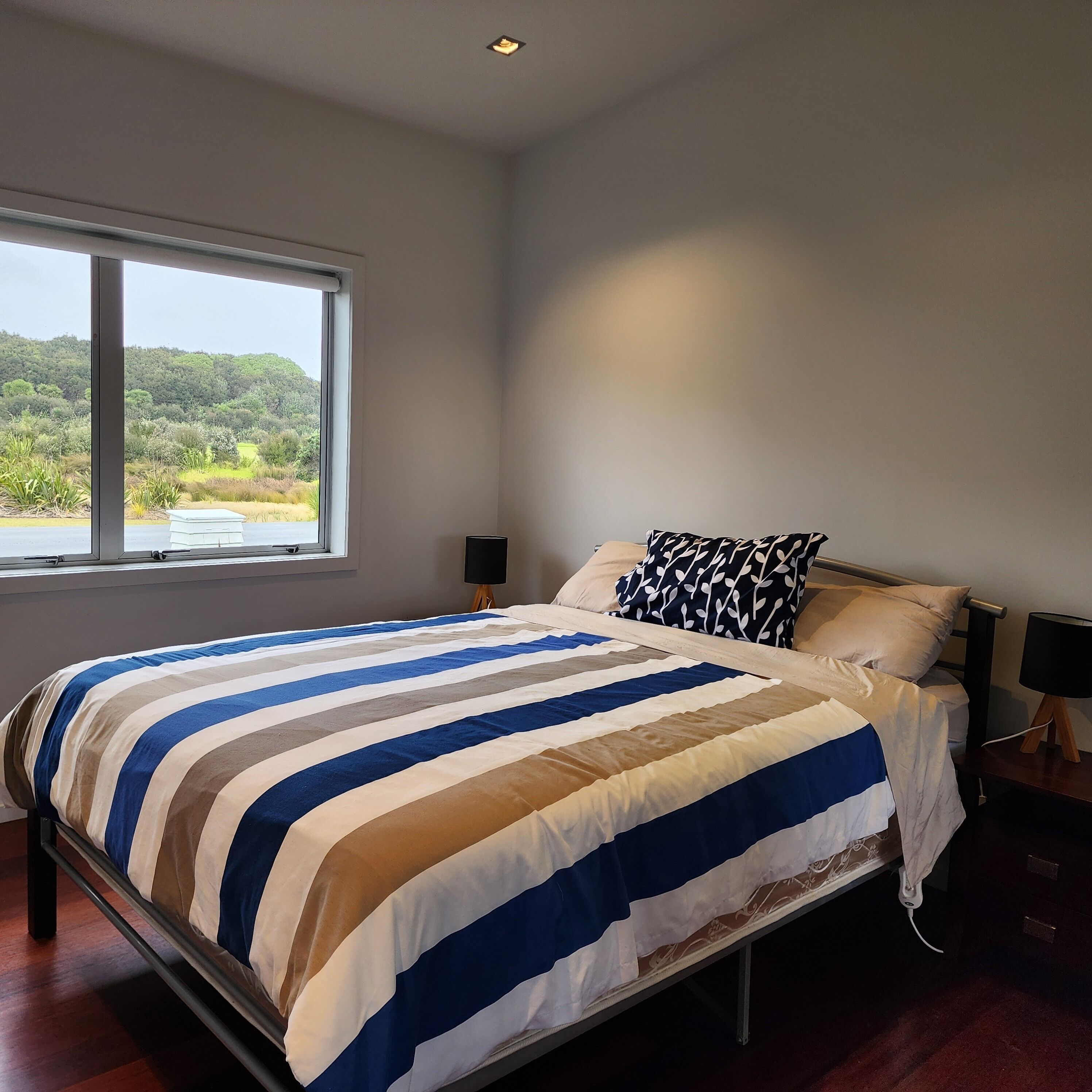 A bedroom with a large bed, a window with a view of greenery, a bedside table with a lamp, and a wall-mounted speaker.