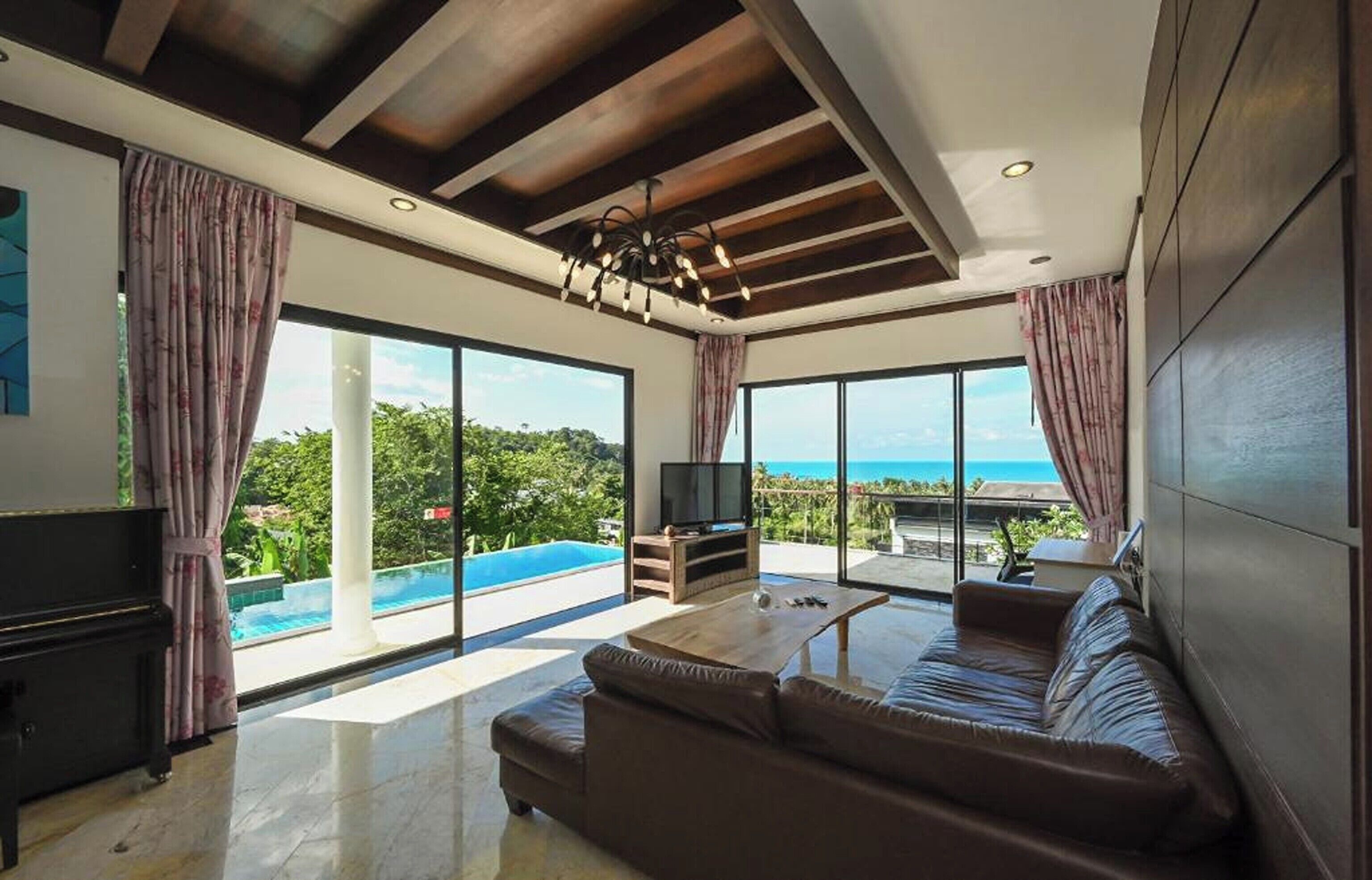 A modern living room with a large sofa, a television, and a view of a swimming pool and ocean.