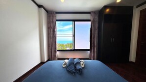 A hotel room with a bed, a window with curtains, a view of the ocean, and a dark door. - Bangpor Seaview Nice Villa (Koh Samui)