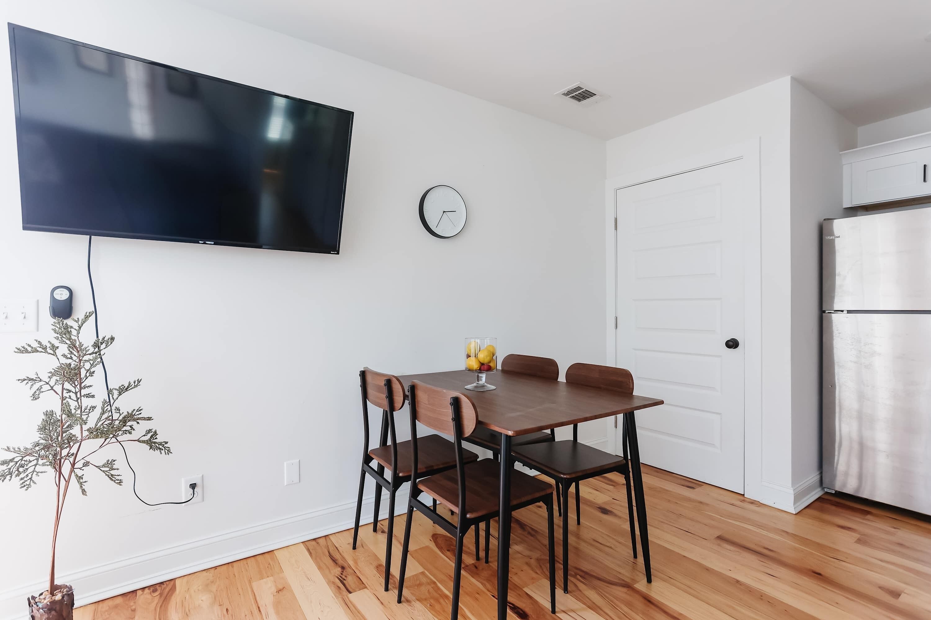 A modern kitchen with a built-in refrigerator, a dining table with chairs, a wall-mounted TV, and a clock.