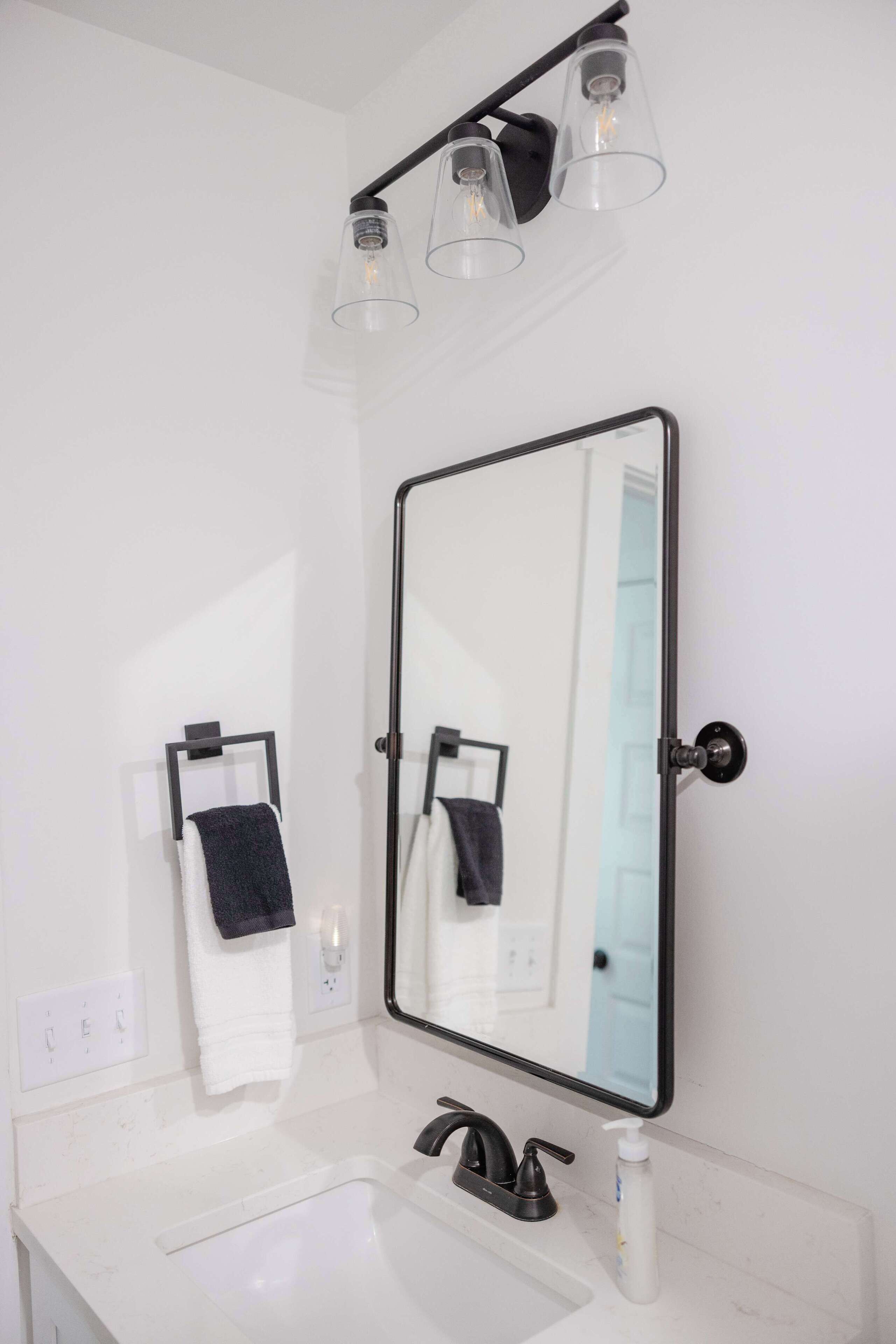 A bathroom with a white sink, black faucet, and a mirror with a black frame. There are two towels hanging on hooks, and a light fixture above the mirror.