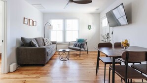 Een moderne woonkamer met een bank, salontafel, eettafel, stoelen, een plafondventilator, een televisie en een raam met zonwering.