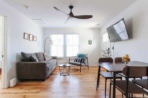 Una sala de estar moderna con un sofá, una mesa de centro, una mesa de comedor, sillas, un ventilador de techo, un televisor y una ventana con persianas.
