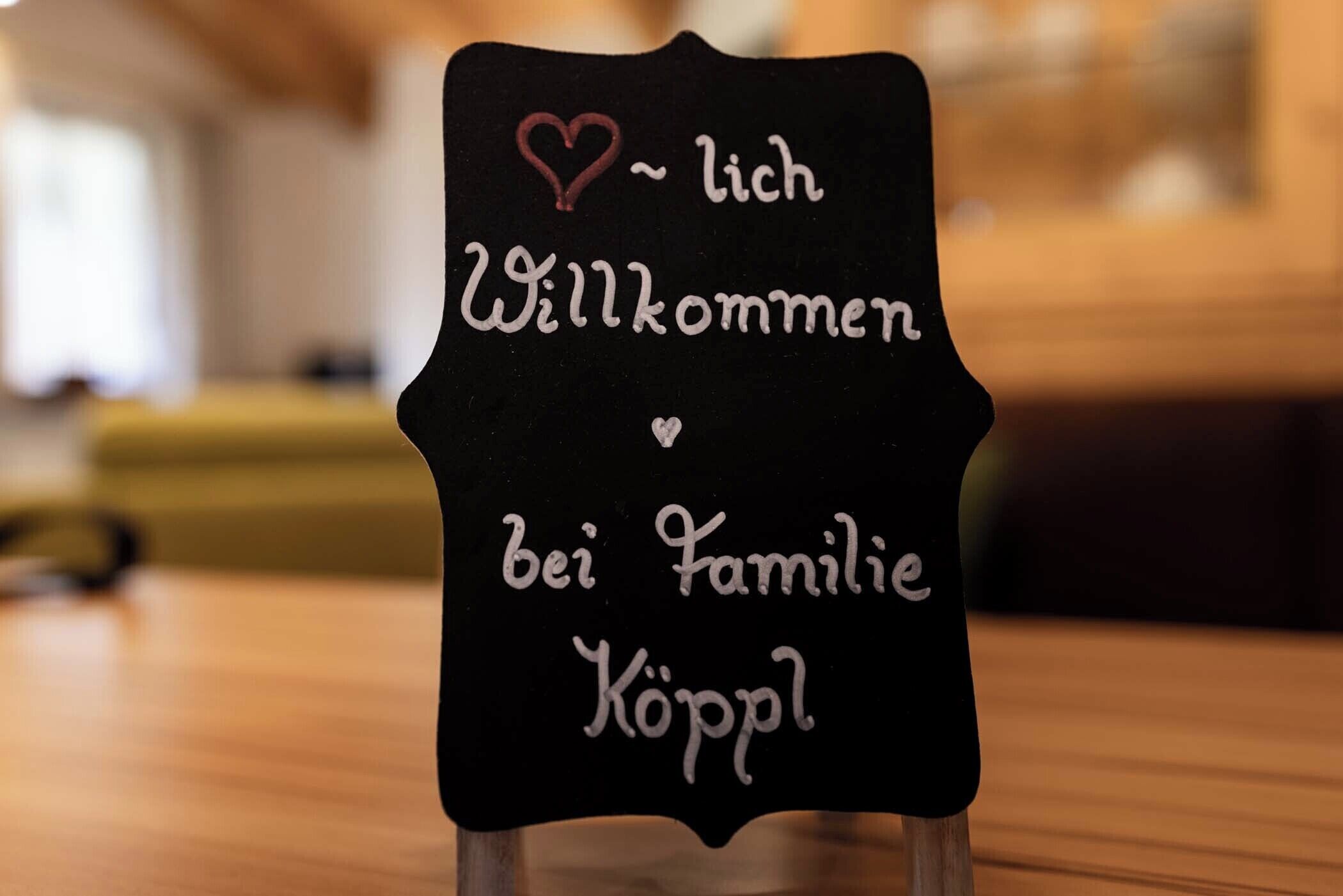Un letrero negro con texto blanco que dice 'Willkommen bei Familie Köppel' y un símbolo de corazón.