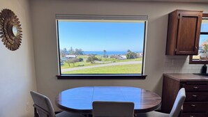 A dining area with a round table, four chairs, a view of a landscape through a large window, and wooden cabinets. - Water View Home Near West Beach (Coupeville)