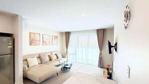 Una sala de estar moderna con un sofá, una mesa de centro y una mesa de comedor. Hay un refrigerador, un reloj en la pared y una ventana con cortinas.