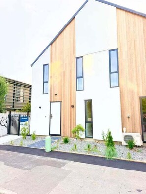 Un bâtiment moderne à deux étages, doté d’une façade blanche et de panneaux en bois décoratifs, d’une entrée sécurisée par un portail et d’un petit jardin.