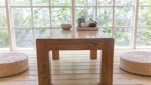 Una mesa de madera con una cesta tejida a cada lado, una bandeja con tetera, tazas y hervidor encima, y vistas a zonas verdes a través de amplios ventanales.