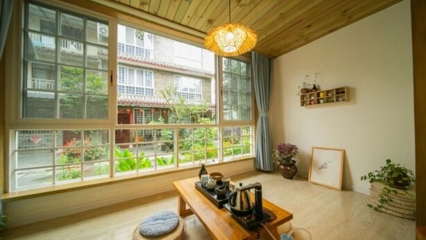 Una habitación con ventanas amplias, techo de madera, una mesa con juego de té, un banco y una estantería con adornos.