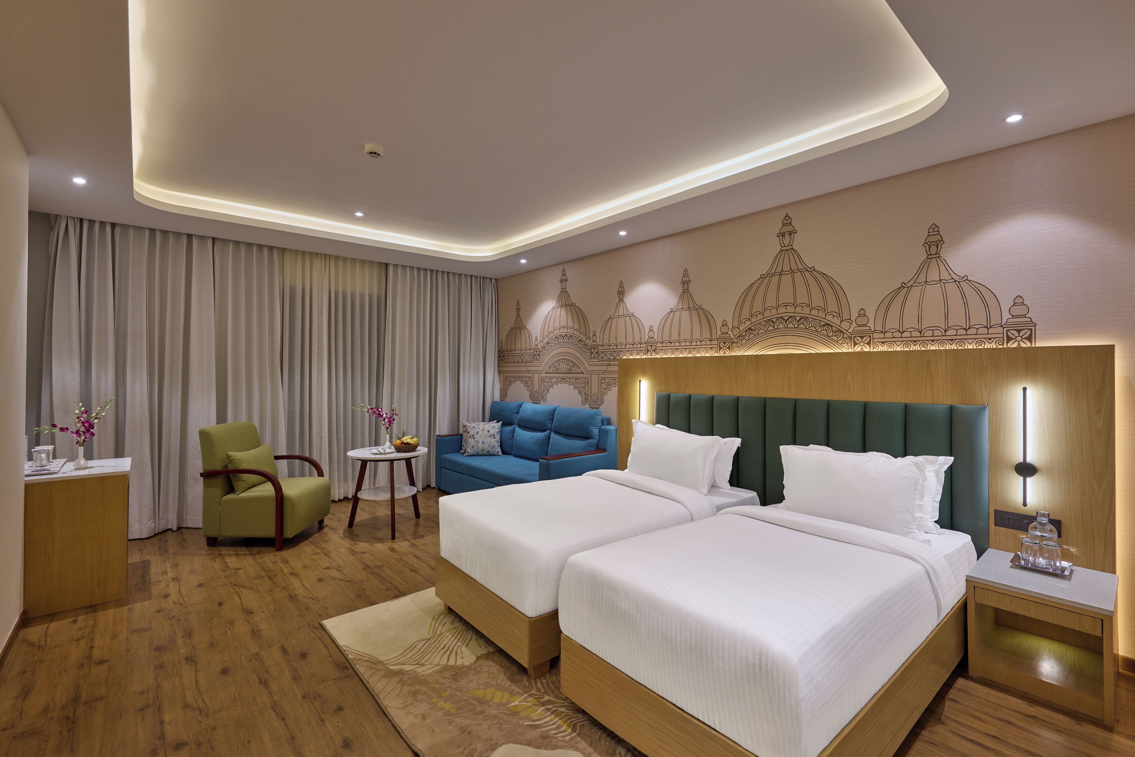 A hotel room with two beds, a blue sofa, a green armchair, a small round table, and a decorative wall mural.