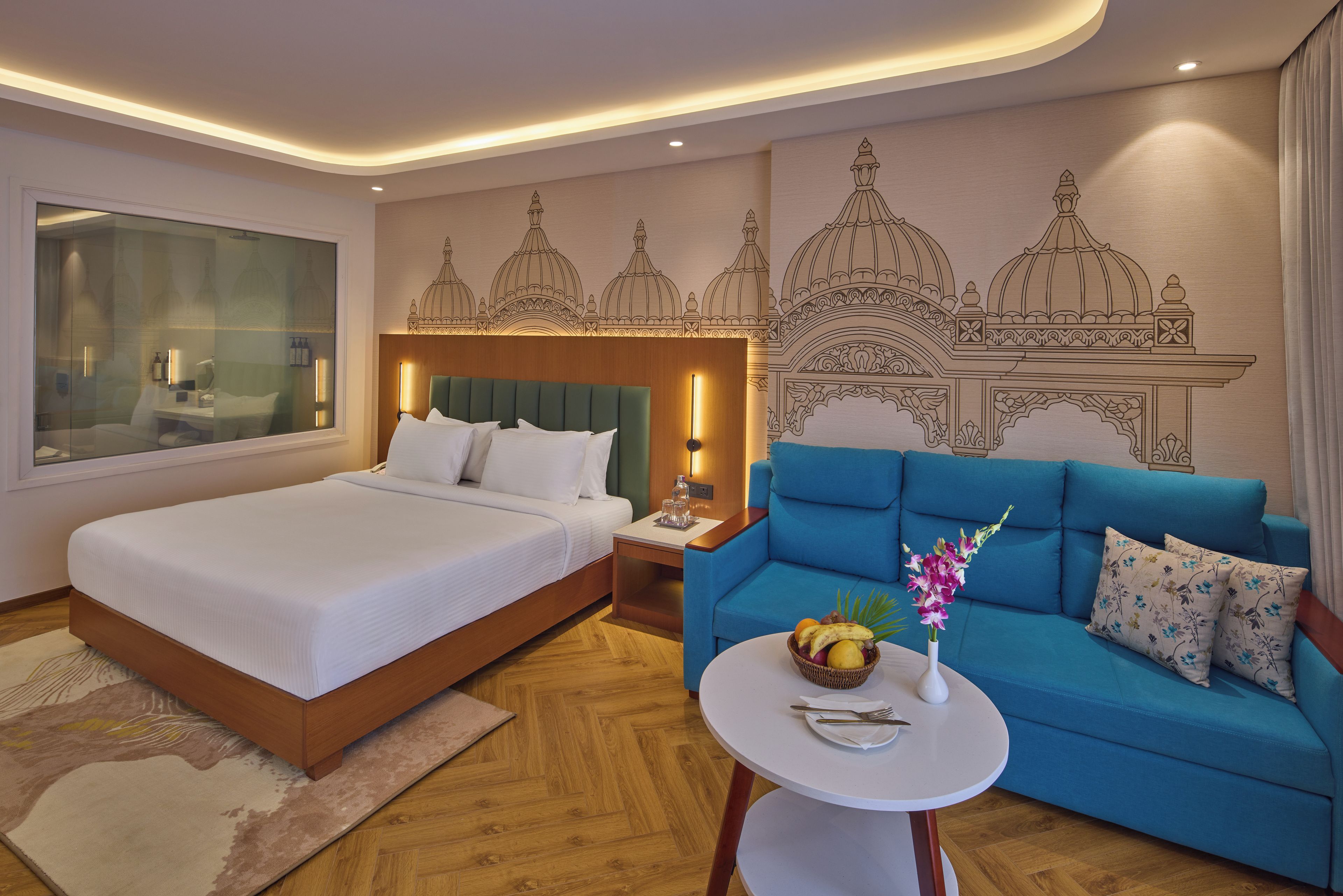 A hotel room with a bed, a blue sofa, a small round table with a vase of flowers, and a wall with a decorative mural.