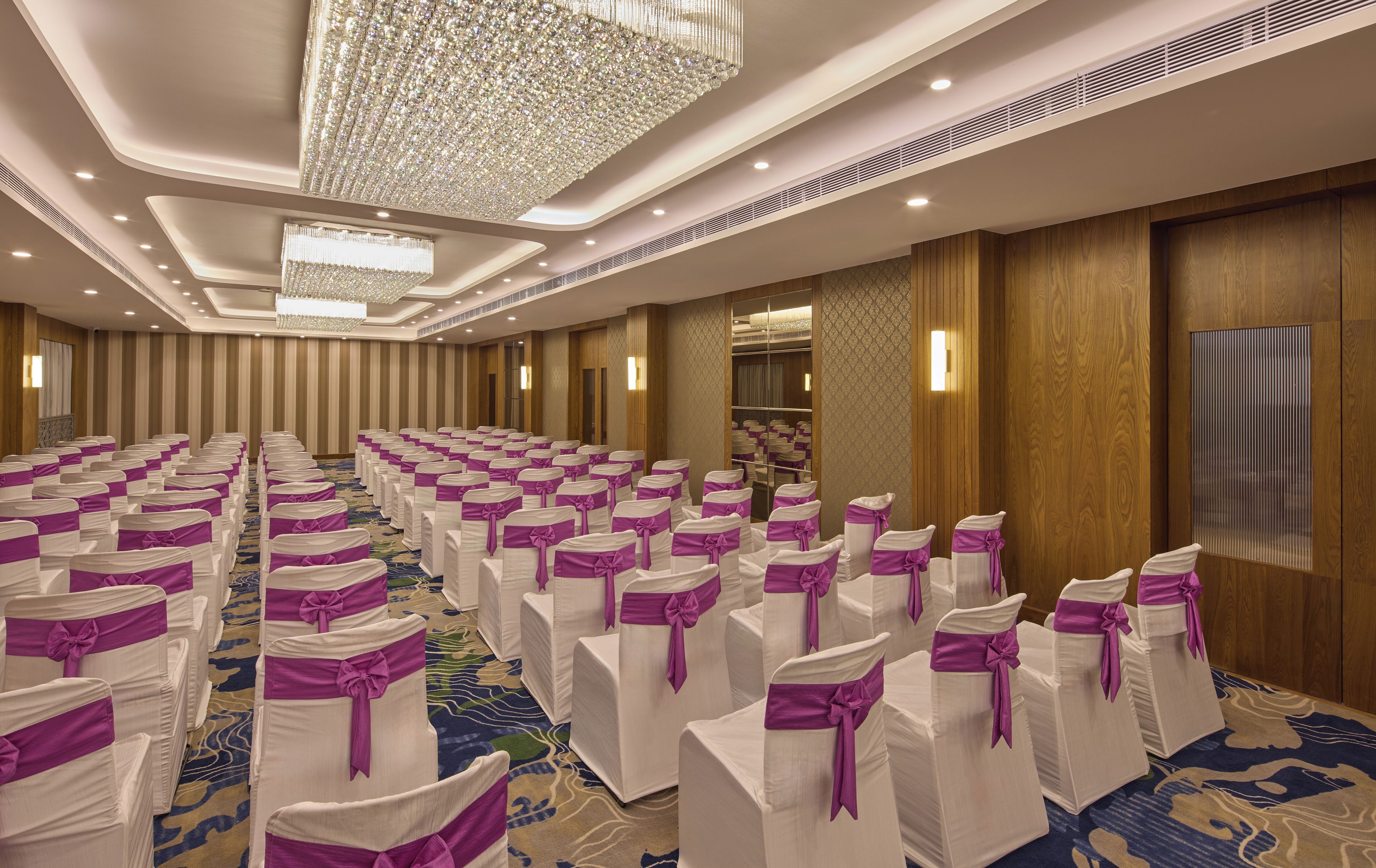 Sala de conferência com fileiras de cadeiras, um lustre grande e um tapete estampado.