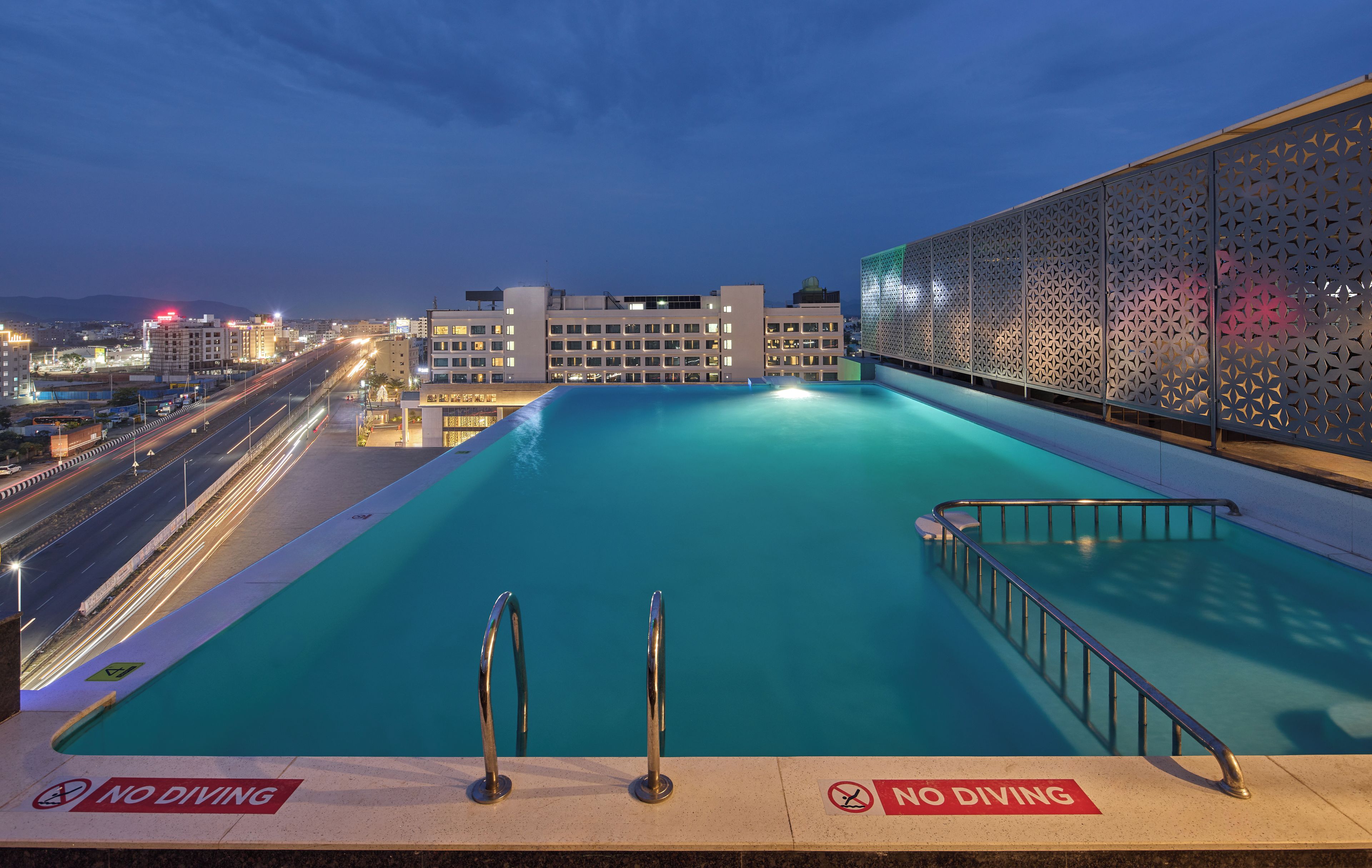 A rooftop swimming pool with a 'No Diving' sign, surrounded by modern buildings and a cityscape at dusk.