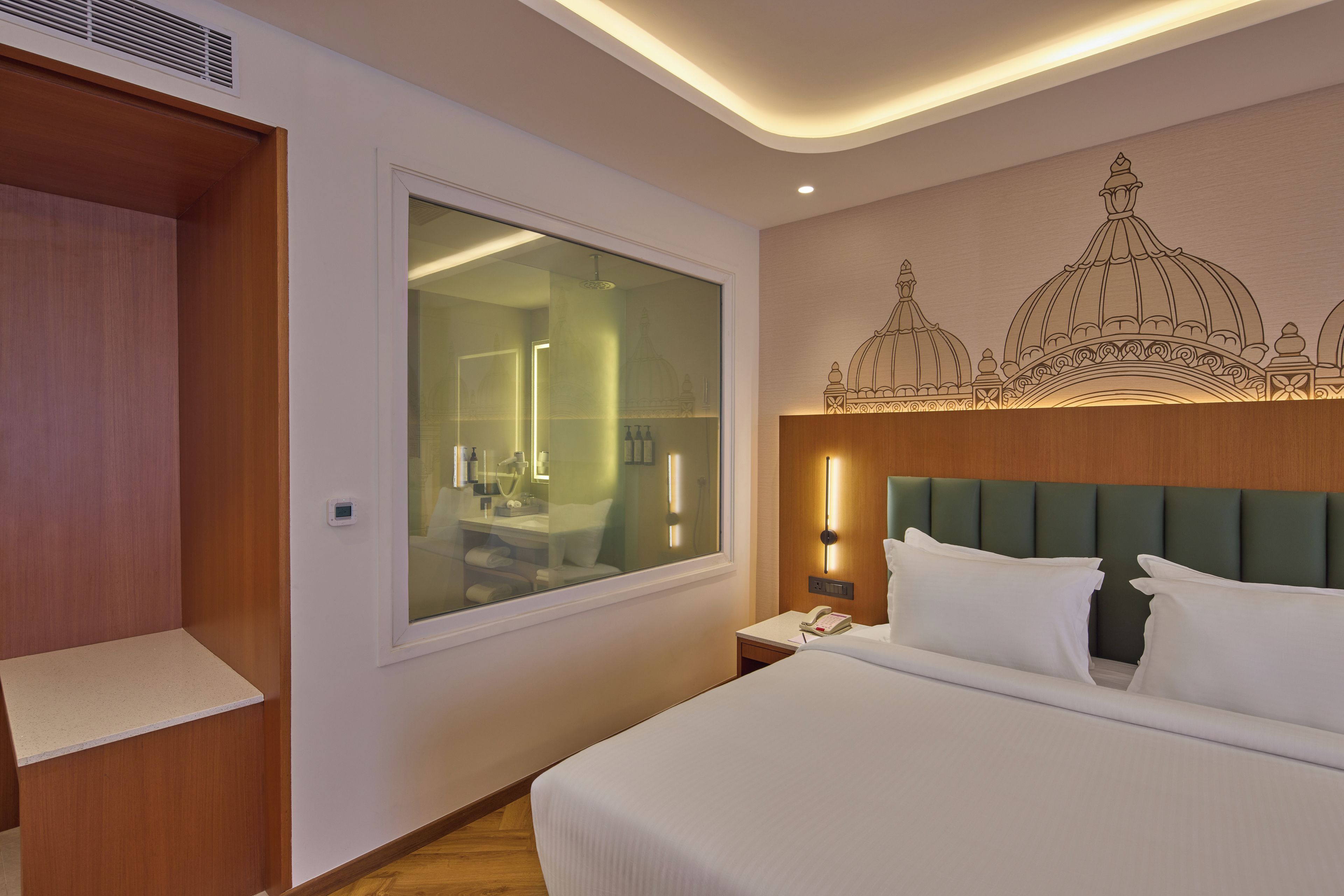 A hotel room with a large bed, a bedside table with a lamp, a wall-mounted phone, and a window with a view of a building.