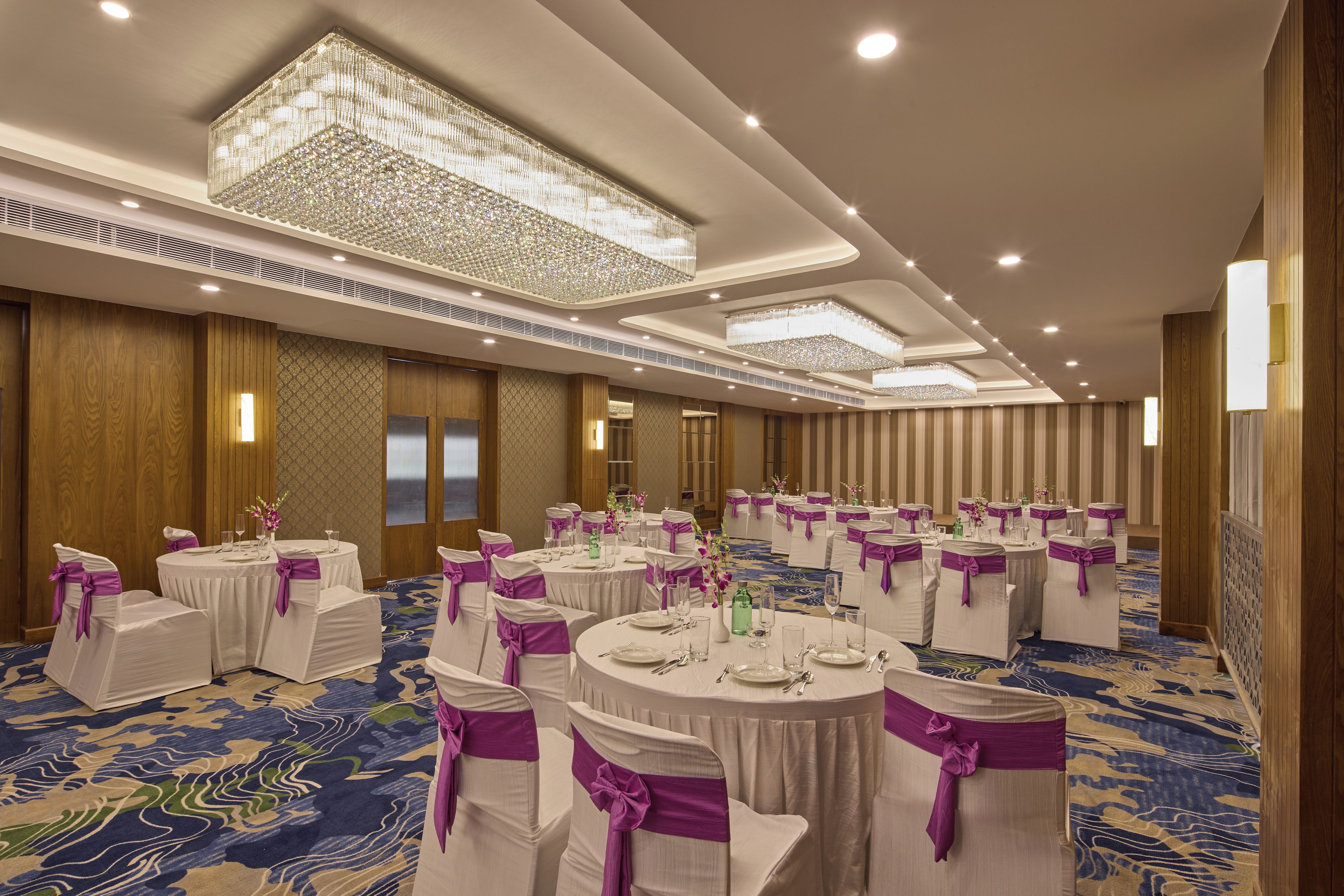 Une salle de banquet avec des tables rondes, des chaises blanches ornées de rubans violets et des lustres.