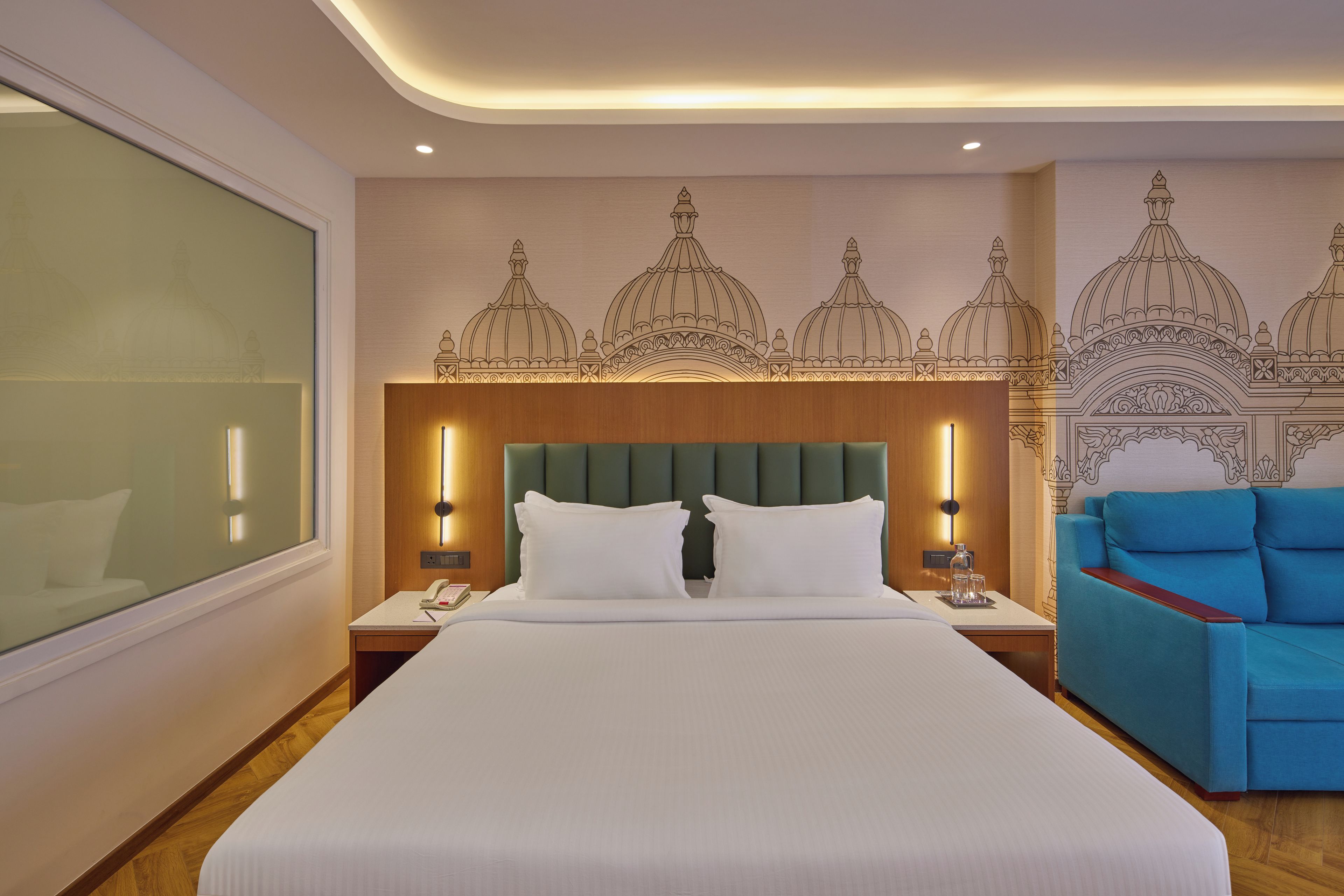 Une chambre d’hôtel avec un grand lit, une tête de lit verte recouverte de tissu, un canapé bleu et des œuvres d’art décoratives au mur.