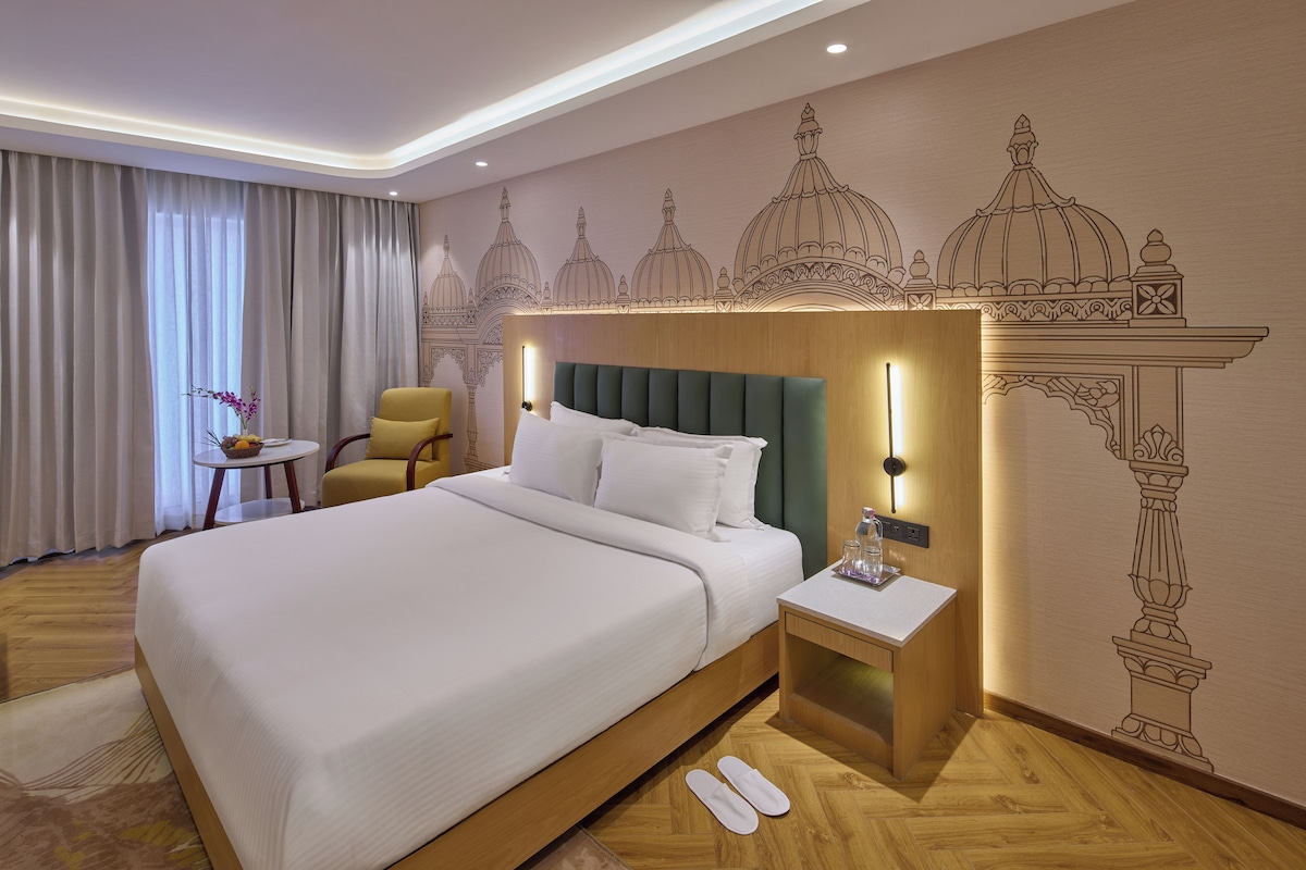 A hotel room with a large bed, a bedside table with a lamp, a chair, and a decorative wall mural.