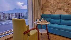 Una habitación con sofá, una silla, una mesita con una bandeja de objetos, una ventana que ofrece vistas a edificios y montañas, y una pared con un diseño decorativo.