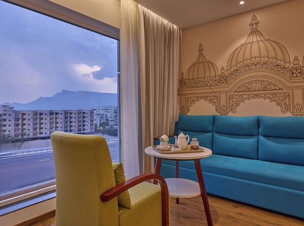 Una habitación con sofá, una silla, una mesita con una bandeja de objetos, una ventana que ofrece vistas a edificios y montañas, y una pared con un diseño decorativo.