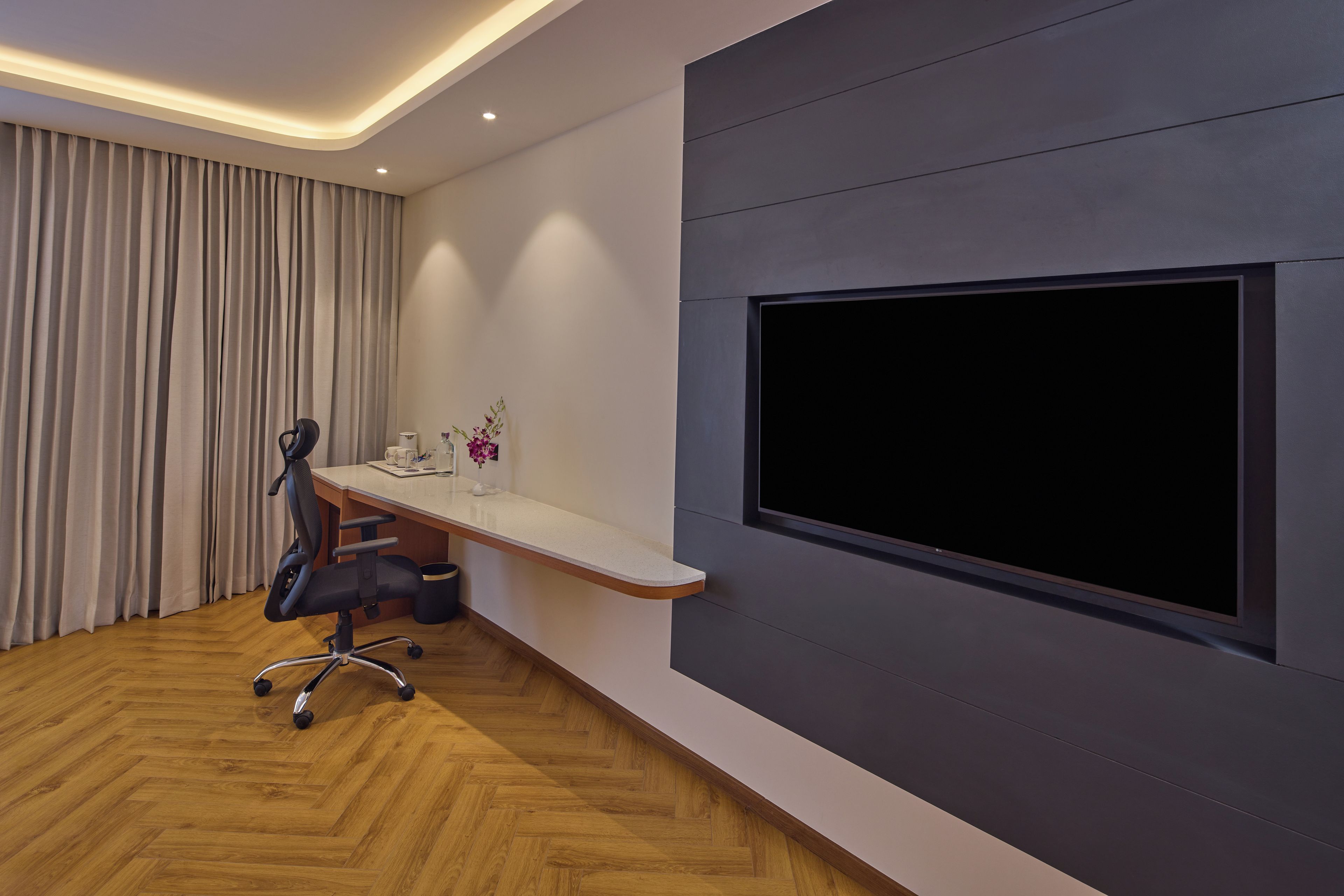 Ein moderner Büroraum mit einem großen Flachbildfernseher an der Wand, einem Schreibtisch mit Stuhl und einem Holzboden mit Fischgrätmuster.