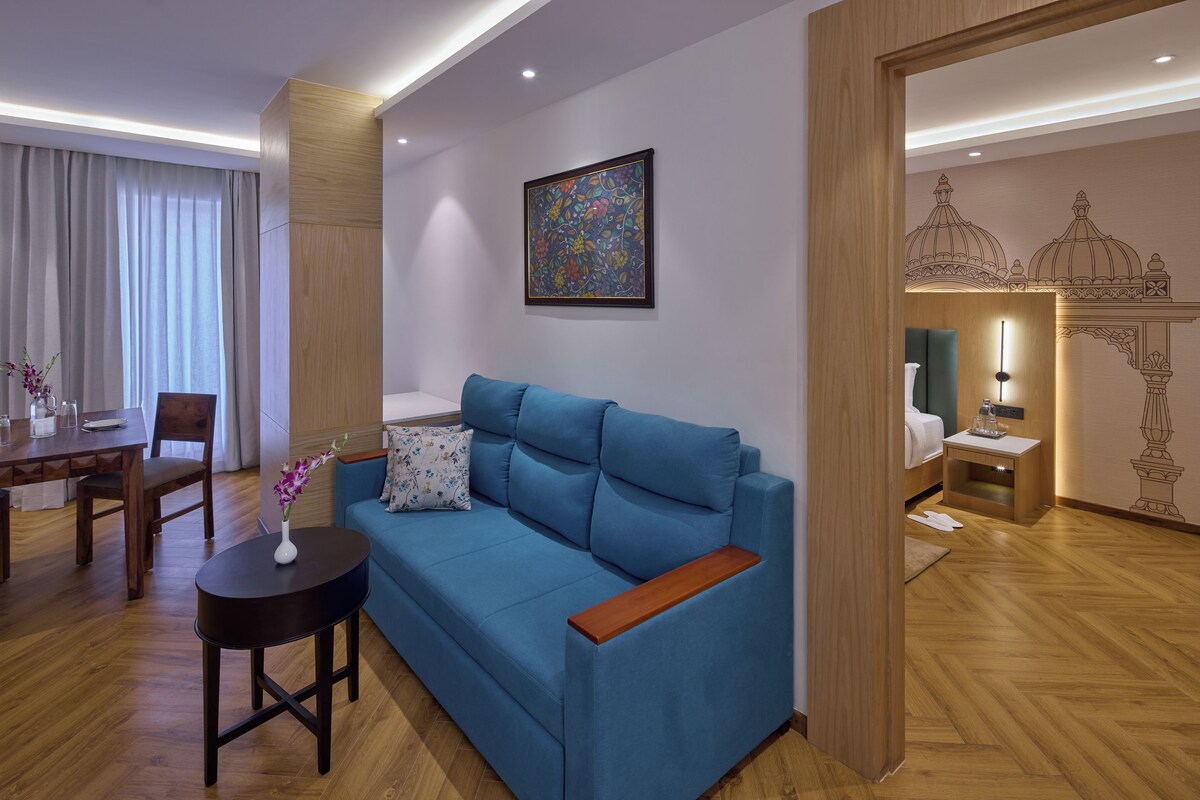 A modern living room with a blue sofa, a small round table with a vase, a dining area with a wooden table and chairs, and a decorative wall piece.