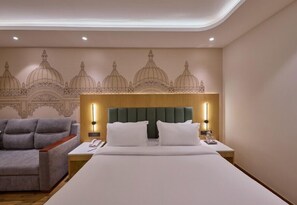 Una habitación de hotel con una cama grande, un cabecero tapizado en verde, dos mesitas de noche con lámparas, un sillón gris y un mural en la pared.