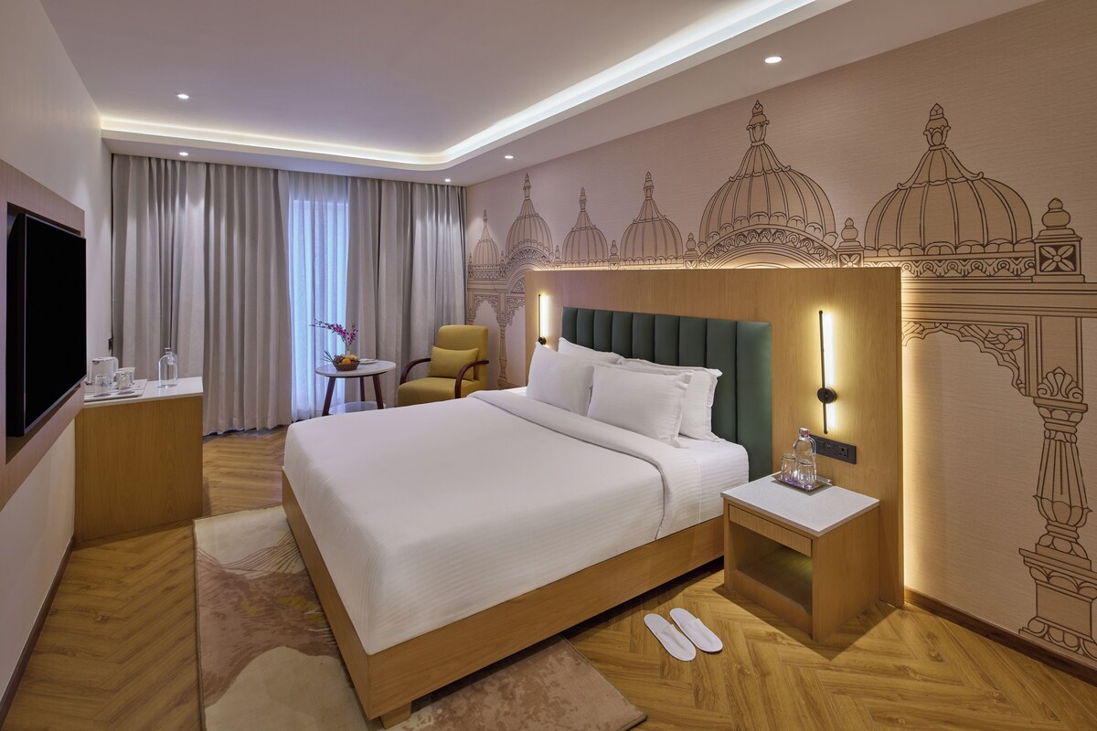 Habitación de hotel con una cama grande, una mesita de noche con lámpara, una silla, un televisor y un detalle decorativo en la pared.