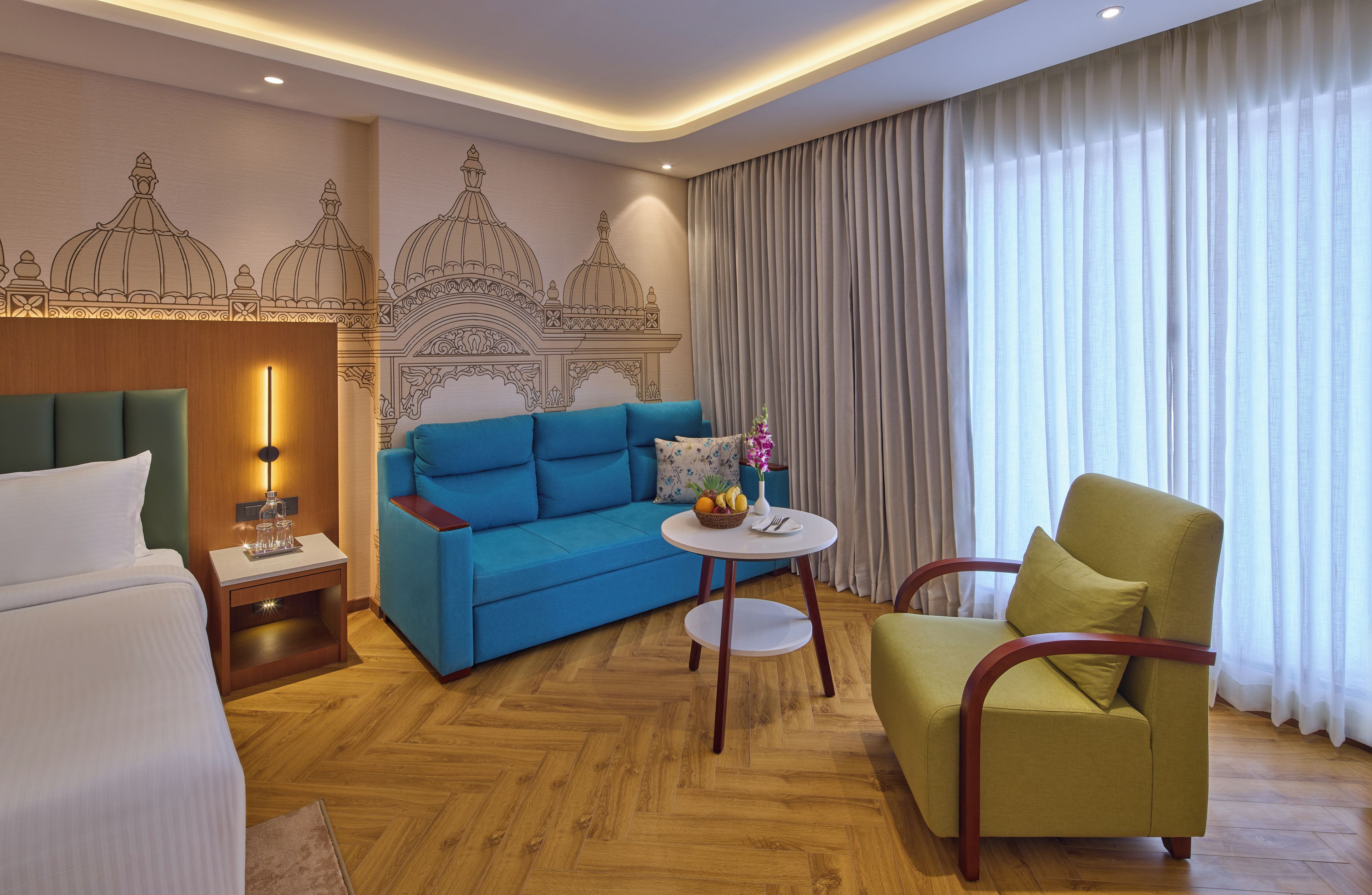 Habitación de hotel con una cama, un sofá azul, un sillón verde, una pequeña mesa redonda con un florero y fruta, y una pared con un diseño decorativo.